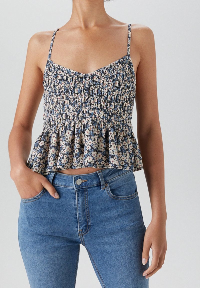 Femme portant un haut smocké sans manches à motifs floraux avec des bretelles spaghetti et un jean taille haute bleu, debout avec une main dans la poche.