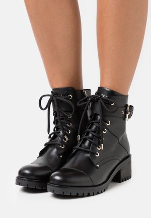Bottines noires en cuir avec lacets à l'avant, sangle à boucle latérale et talon carré, portées sur jambes nues sur fond blanc.