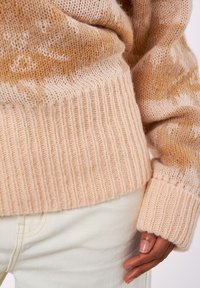 Suéter de punto extragrande hecho de hilo suave y de color beige claro con sutiles patrones naranjas y puños acanalados, combinado con pantalones de color crema.