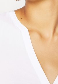 Blusa bianca con scollo a V, realizzata in tessuto morbido, caratterizzata da un design pulito. Tono della pelle visibile al collo senza ulteriori dettagli.