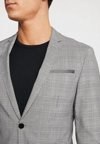 Man med grå rutigt blazers över en svart t-shirt med rund hals, som visar blazerns slag, bröstficka och detaljer på knapparna.