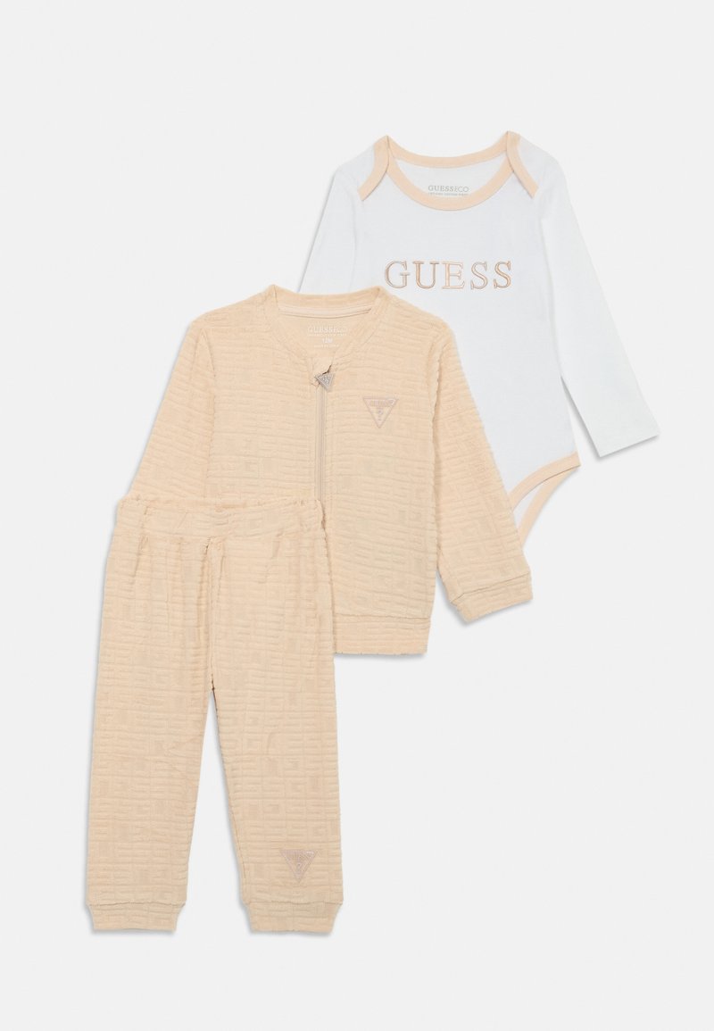 Beigu krāsas frotē auduma jaka ar teksturētu rakstu, saderīgas bikses un baltu garām piedurknēm bodiju ar "GUESS" logotipu maigi rozā krāsā.