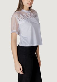 Camiseta blanca recortada con mangas de malla transparente y patrón geométrico bordado en el cuello, combinada con pantalones negros. Textura suave.