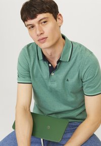 Jeune homme en polo vert et jean bleu tenant une tablette ou un dossier vert, regardant légèrement sur le côté sur un fond uni.