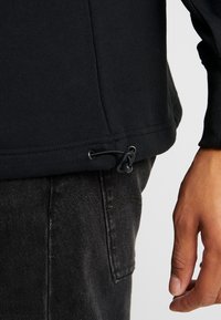 Mano che regola il cordino nero all'orlo di una maglietta nera a maniche lunghe, con jeans di denim scuro visibili.