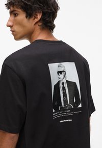 Črna bombažna majica s potiskom na hrbtni strani, ki prikazuje postavo v obleki z sončnimi očali in besedilom spodaj, kar ponuja sodoben dizajn.