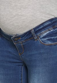 Close-up of a person wearing tight blue jeans with the button undone and a light gray shirt tucked inside.