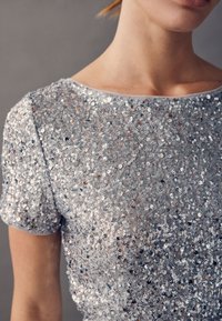 Top in paillettes argento con maniche corte, caratterizzato da una scollatura tonda e una texture liscia, impreziosito da paillettes circolari scintillanti.