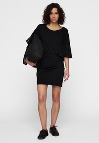 Vestido de algodón negro con una parte superior holgada y una parte inferior ajustada, que presenta fruncidos laterales, complementado con un gran bolso negro y zapatos oscuros.