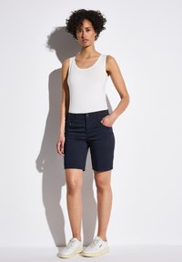 Pantalones cortos azul marino con un diseño ajustado, que cuentan con bolsillos frontales y un cierre de botón. Combinados con una camiseta sin mangas blanca y deportivas claras.