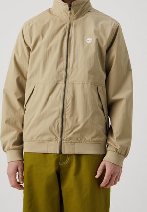 Veste légère beige zippée avec col montant et logo sur la poitrine, portée avec un pantalon vert olive.
