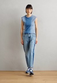 Világoskék bordás, ujjatlan felső, amely világoskék mosott farmernadrággal van párosítva, sötétebb kék oldalsávval. A modellen sötétkék sportcipő van.