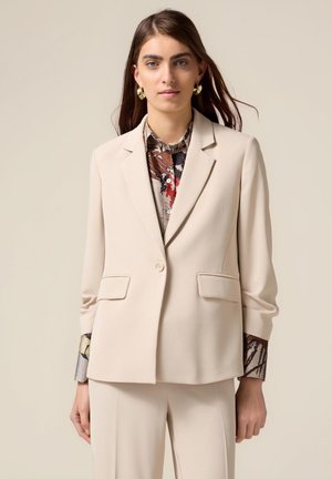 Blazer beige in un tessuto liscio con un solo bottone e due tasche frontali. Il modello indossa una camicia a fantasia con le maniche arrotolate.