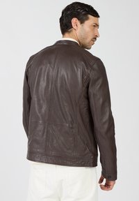 Brązowa skórzana kurtka o gładkiej fakturze, z prostym kołnierzem i widocznymi szwami. Tył pokazuje dopasowany krój i minimalne zdobienia.