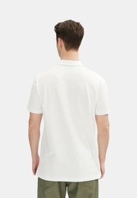 Polo shirt bianco a maniche corte con colletto classico, texture liscia e orlo dritto. La vista dal retro mostra un design semplice senza decorazioni.