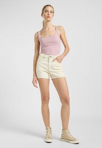 Vaaleanpunainen ribbineuleinen hihaton paita yhdistettynä korkeavyötäröisiin vaaleisiin shortseihin. Mallilla on päällään beige korkeavyötäröiset lenkkitossut. Tausta on yksinkertainen valkoinen.