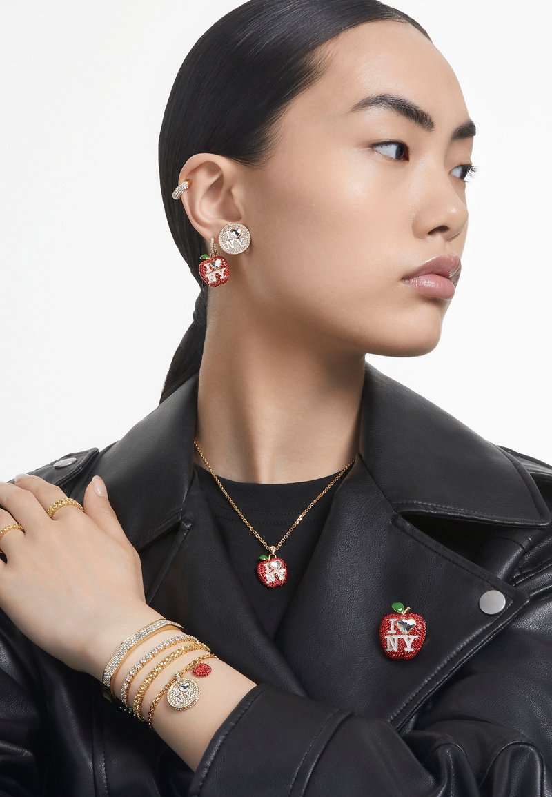 Brincos em forma de maça vermelha e um colar com acentos "I ♥ NY", pulseiras de corrente dourada e uma jaqueta de couro preta. Texturas brilhantes por toda a parte.