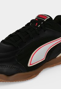 Μαύρο παπούτσι Puma με λευκή και κόκκινη πλαϊνή ρίγα, μαύρα κορδόνια, καουτσούκ σόλα και λογότυπο Puma στη γλώσσα πάνω σε ανοιχτό φόντο.