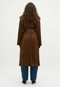 Bruine, lange trenchcoat met een strik riem, gemaakt van zacht materiaal. Heeft schouderepauletten en een rugventilatie, gecombineerd met losse blauwe jeans.