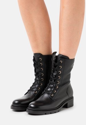 Botas de combate de cuero negro hasta la mitad de la pantorrilla con cordones, cremalleras laterales y tacones bajos gruesos, usadas sobre piernas desnudas contra un fondo sencillo.