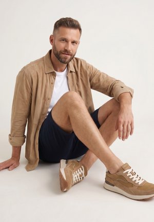 Bärtiger Mann sitzt auf dem Boden, trägt ein beiges Hemd, weißes T-Shirt, marineblaue Shorts und beige Turnschuhe, posiert vor einem einfarbigen Hintergrund.