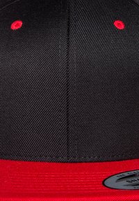 Cappellino nero e rosso con una parte superiore nera testurizzata, visiera rossa liscia e un'etichetta rotonda con logo argentato sul lato. Cuciture rosse visibili.