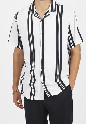 Mann trägt ein weißes kurzärmliges Hemd mit vertikalen schwarzen Streifen, kombiniert mit schwarzen Hosen und einem silbernen Armband.