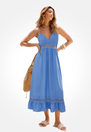 Kobieta w niebieskiej sukience midi bez rękawów z koronkowymi detalami, niosąca plecioną torbę, ozdobiona bransoletkami i warstwowymi naszyjnikami.