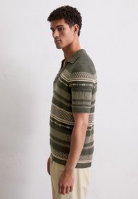 Gebreide polo in donkergroen met beige en witte geometrische patronen, voorzien van een kraag en korte mouwen, gecombineerd met lichte broek.