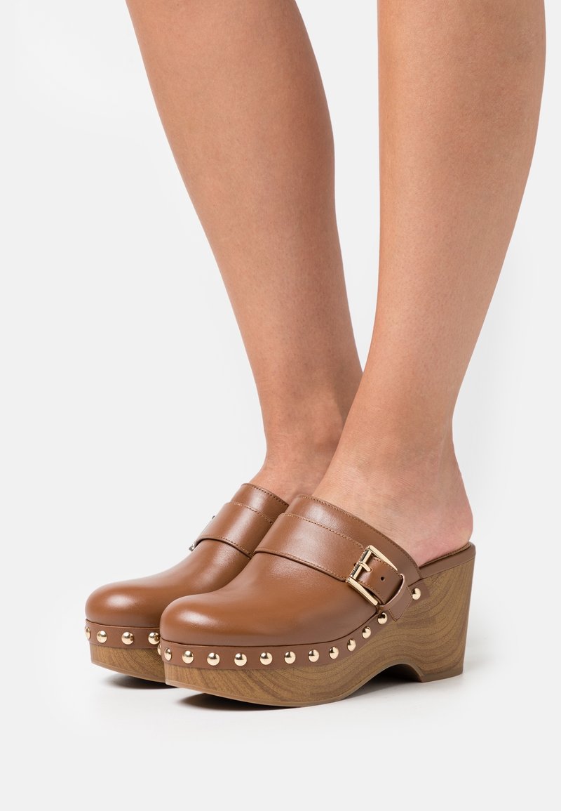 Sabots en cuir marron à bout fermé, avec une large sangle et une boucle en accent. Talon compensé aspect bois orné de clous métalliques.