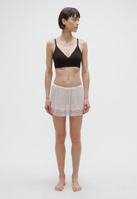 Črna bralette majica s tankimi naramnicami, združena z belimi teksturiranimi kratkimi hlačami z čipkastim robom. Model stoji bos na enostavni podlagi.