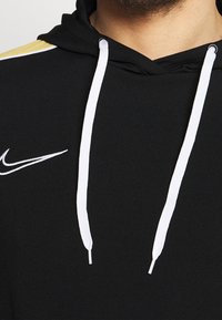 Czarna bluza z kapturem wykonana z miękkiego materiału z białymi sznurkami. Posiada białe logo Nike na klatce piersiowej oraz złoty akcent na ramieniu.