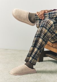 Uzavreté béžové papuče s rybičkovým vzorom, nosené s hnedými ponožkami a károvanými nohavicami, umiestnené na textúrovanej svetlej koberci.