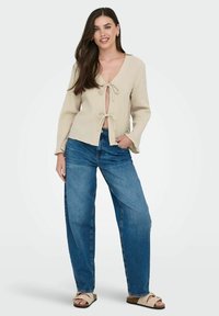 Beige Bluse mit langen Ärmeln und Schleifenverschluss, tiefem V-Ausschnitt und strukturiertem Stoff; kombiniert mit lockeren, blauen Denim-Jeans und braunen Riemchensandalen.