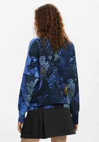 Pullover blu lavorato a maglia con motivi floreali e geometrici, maniche lunghe e orlo a coste. Indossato con una gonna nera.