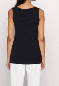 Donna che indossa un top nero senza maniche e pantaloni bianchi, vista di spalle su uno sfondo chiaro e semplice.