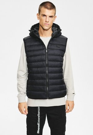 Czarna pikowana kamizelka z kapturem, z przodu na zamek błyskawiczny i regulowanym ściągaczem. Noszona na kremowej koszuli z długim rękawem.