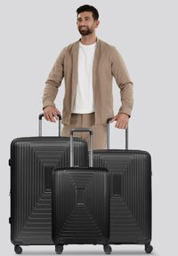 Uomo in giacca e pantaloni beige che tiene le maniglie di tre valigie rigide nere con manici telescopici su uno sfondo grigio.
