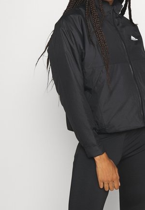 Personne portant une veste zippée Adidas noire et un pantalon noir, debout devant un fond blanc uni.