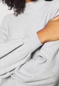 Pull gris en maille avec un col montant et des manches volumineuses aux poignets côtelés, mettant en valeur une texture douce et un design décontracté.