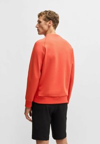 Oranges Sweatshirt mit Rundhalsausschnitt, gerippten Bündchen und Saum. Kombiniert mit schwarzen Shorts wirkt der Stoff glatt und leicht strukturiert.