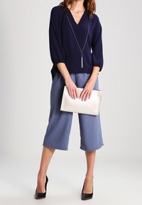 Blouse bleu marine à col en V et manches bouffantes, associée à un pantalon culotte bleu clair. Une pochette métallique et des talons hauts noirs complètent la tenue.