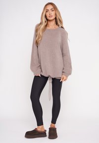 Jasnobrązowy, oversize'owy sweter wykonany z dzianiny, z długimi rękawami i ściąganym dołem, zestawiony z czarnymi legginsami oraz ciemnobrązowymi klapkami z czerwonymi akcentami.