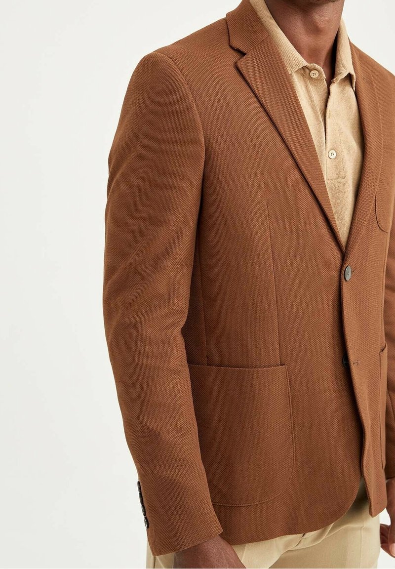 Bruine getextureerde blazer met een enkele knoop, ingesneden revers en opgestikte zakken. Draagt over een beige overhemd met kraag. Glad stoffen oppervlak.