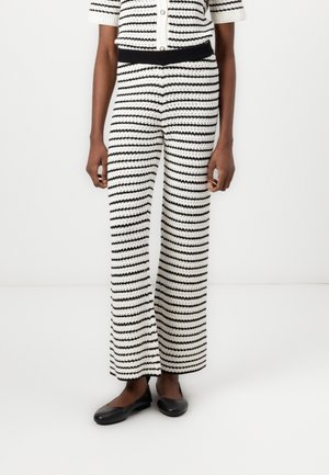 Frau trägt hoch taillierte, weit geschnittene, schwarz-weiß gestreifte Häkelhosen mit schwarzen Ballerinas, steht vor weißem Hintergrund.