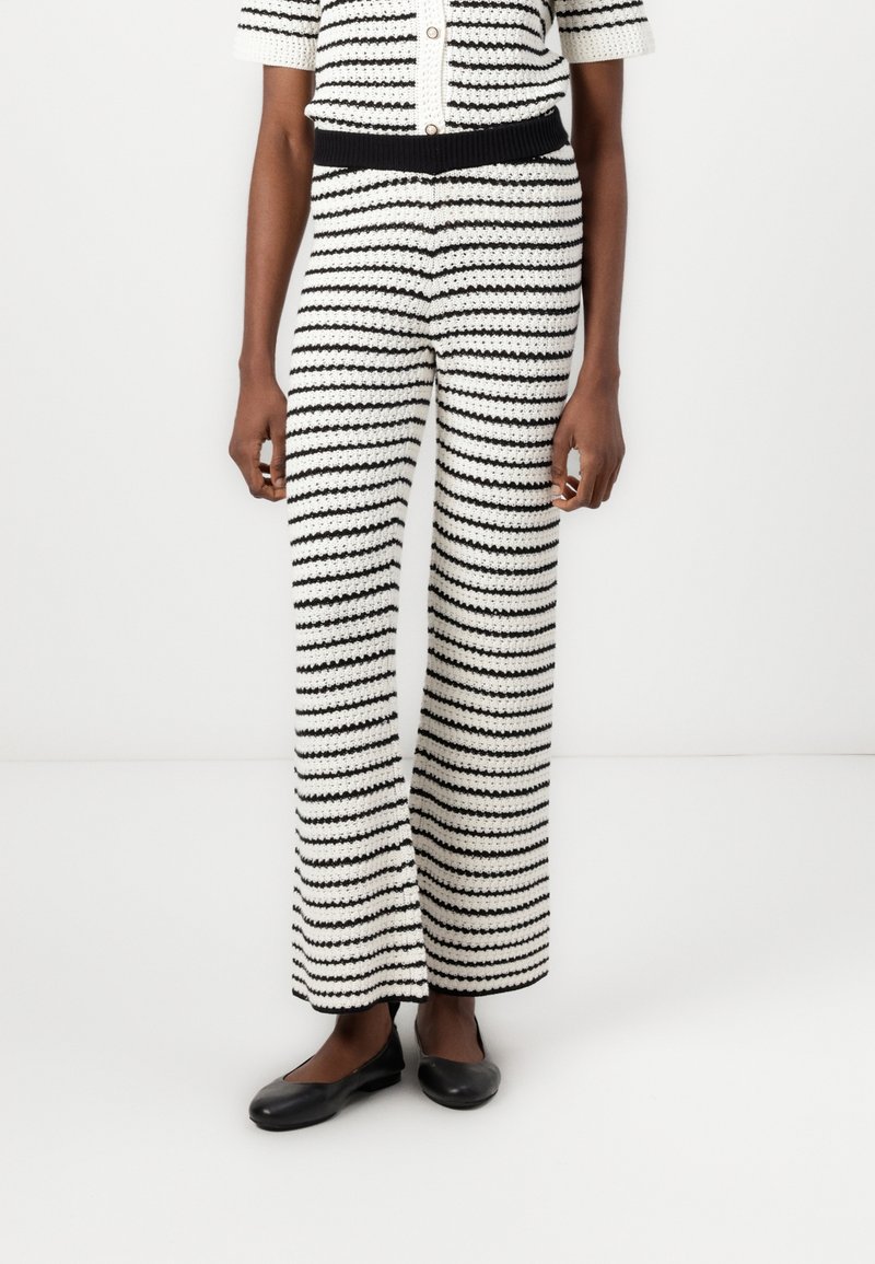 Femme portant un pantalon crochet à rayures noires et blanches, taille haute et jambes larges, avec des ballerines noires, debout devant un fond blanc.