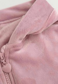 Różowy polarowy ubiór o miękkiej strukturze, z zamkiem błyskawicznym zakończonym suwakiem w kształcie serca oraz z kołnierzem zapinanym na złożenie.