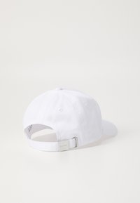 Hvit caps med buet skjerm, justerbar stropp bak, og et lite metallisk logo merke. Laget av stoff med en glatt tekstur.