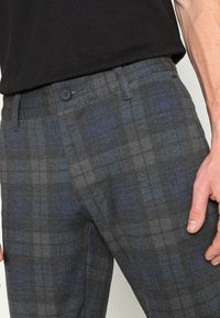 Ruitbroek in donkergrijs met blauwe accenten, voorzien van een knoopsluiting en een lichte glans. Het materiaal lijkt gestructureerd en is geschikt voor casual kleding.