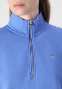 Lichtblauwe rits sweatshirt met hoge kraag, voorzien van een zilveren rits en een klein geborduurd logo op de linkerborst. Zachte textuur.